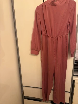 Rosa långärmad jumpsuit - En enkel och snygg rosa jumpsuit med långa ärmar och resår i midjan. Modellen har hög hals och raka ben. Perfekt för dig som gillar en avslappnad men ändå trendig stil. Materialet känns mjukt och bekvämt mot huden.