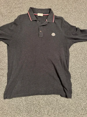 Mörkgrå långärmad pikétröja från Moncler - Snygg mörkgrå pikétröja från Moncler med klassisk krage och knappar framtill. Tröjan har Moncler-logga på bröstet och rödvit randig detalj på kragen. Perfekt för en stilren och avslappnad look.