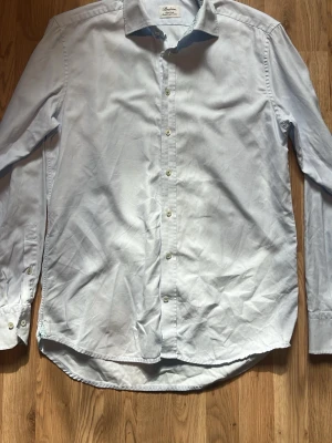 Ljusblå skjorta från Stenströms - Snygg ljusblå skjorta från Stenströms med diskret rutigt mönster och gröna detaljer på insidan av kragen. Skjortan har klassisk krage, knappar framtill och är långärmad. Tillverkad i mjuk bomull för en bekväm känsla. OBS! Har en liten fläck vid magen inget man märker.