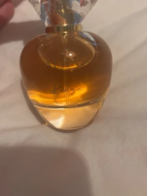 Alani Kenza Eau de Parfum - Elegant parfym från Alani med namnet Kenza. Flaskan är rundad och genomskinlig med gyllene-orange vätska och ett dekorativt, kristalliknande lock. Texten på flaskan är i guld. Perfekt för dig som gillar lyxiga dofter och snygg design på parfymflaskan.