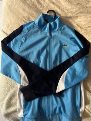Blå träningsjacka från Lacoste - Snygg blå träningsjacka från Lacoste med dragkedja framtill. Jackan har mörkblå och vita detaljer längs ärmarna och sidorna samt den klassiska Lacoste-loggan på bröstet. 