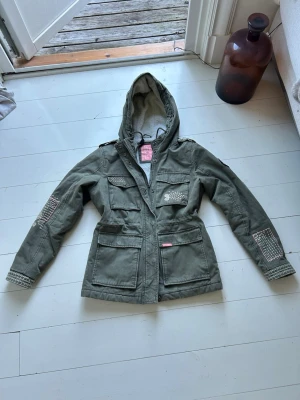 Mörkgrön superdry jacka - Väldigt fin superdry jacka. Den har vita och rosa broderier vilket gör den väldigt unik. Säljes för den är för liten. Storlek 38 (M). Tveka inte att höra av er vid frågor!