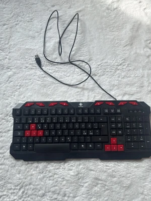 Svart och röd gamingtangentbord USB - Gamingtangentbord med USB-anslutning i svart plast med röda detaljer på piltangenter, WASD och funktionsknappar. Fullstort tangentbord med numerisk del och tydliga knappar, perfekt för gaming eller datoranvändning.
