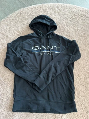 Svart hoodie från GANT med tryck - Snygg svart hoodie från GANT med stor logga och texten 'EST. 1949' på bröstet. Klassisk huva med dragsko och ribbade muddar vid ärmar och nederkant. Tillverkad i mjukt bomullsmaterial som är skönt att ha på sig. Perfekt för en avslappnad och stilren look.