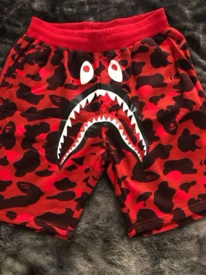 Röda BAPE shark camo shorts - Säljer ett par röda BAPE shorts med ikoniskt shark-motiv framtill och svart-röd camouflage-mönster. Elastisk midja och cool design som sticker ut. Perfekt för dig som gillar streetwear och vill ha något unikt.