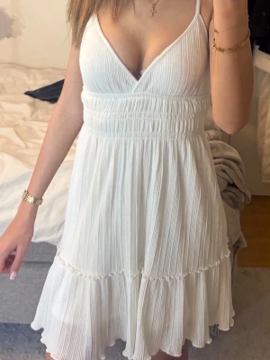 Vit söt sommarklänning med volang - Säljer en vit kortklänning med smala axelband och v-ringning. Klänningen har volangdetaljer, korsade axelband i ryggen och är gjord i ett lätt, plisserat material som passar perfekt till sommaren. Sitter snyggt i midjan och har en luftig passform.