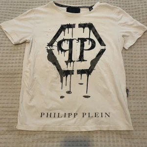 Vit Philipp Plein t-shirt med tryck - Snygg vit t-shirt från Philipp Plein med stort svart grafiskt tryck och logga framtill. Klassisk passform med korta ärmar och rund halsringning. Perfekt för dig som gillar streetwear och vill sticka ut med en exklusiv look. Litet hål i nacken(visas i bilden) därför det billiga priset.