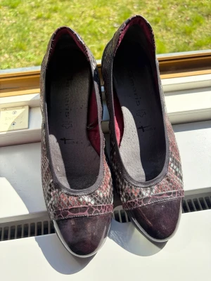 Mönstrade ballerinaskor från Tamaris - Säljer ett par ballerinaskor från Tamaris med snyggt ormmönster i vinrött, svart och grått. Skorna har rund tå, svarta detaljer i mockaimitation och en bekväm gummisula. Perfekta för dig som vill ha något unikt och trendigt till vardagslooken.