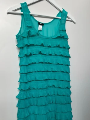 Mini klänning strl 34/36 - En söt, kort turkos klänning från H&M med många lager av plisserade volanger. Den har en rund halsringning och är tillverkad i ett tunt chiffonglikt material med ett underliggande foder.   Strl 34 men passar även 36