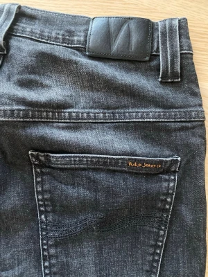 Svart/grå jeans från Nudie Jeans - Snygga svarta jeans från Nudie Jeans med klassisk femficksmodell och diskret logga på bakfickan. Jeansen har raka ben och är tillverkade i slitstark denim med en cool tvättad look. Perfekta för dig som gillar stilrena och tidlösa jeans.