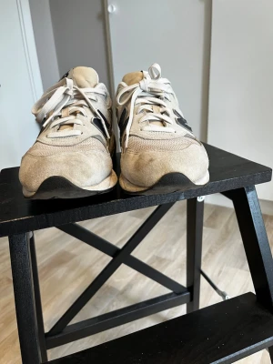 Beige New Balance 580 sneakers - Snygga New Balance 580 sneakers i beige mocka och mesh med vita snören och svarta detaljer. Klassisk chunky sula och ikoniskt N på sidan. Perfekta för dig som gillar streetwear och vill ha en stilren men sportig look.
