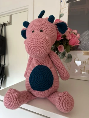 Virkad rosa drake  - Söt virkad rosa drake med petroleumblå detaljer i akrylgarn. Även denna har extra vikt i kroppen så den sitter stadigt. Den väger ungefär 850g och är ca 35 cm hög. Den har säkerhetsögon som inte rekommenderas för barn under 3 år!🩷