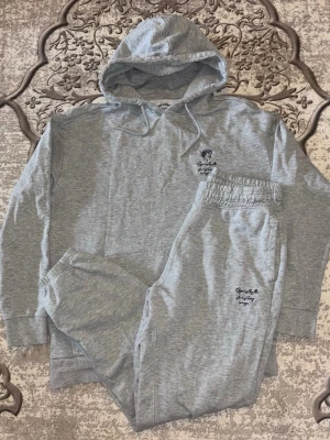 Grått mjukisset med hoodie och byxor - Superbekvämt grått loungewear-set med hoodie och matchande byxor. Hoodien har dragsko och en liten broderad detalj på bröstet, medan byxorna har resår i midjan och broderad text på benet. Perfekt för chill dagar hemma. Använt bara fåtal gånger, nästan ingen tecken på användning och den har bara ligget där. Några frågor bara att ställa. 