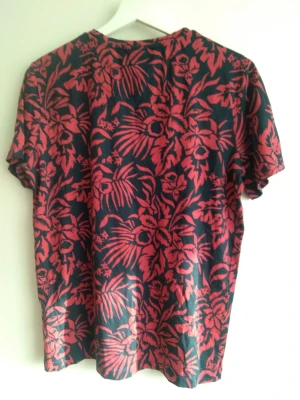 Blommig t-shirt i rött och mörkblått - T-shirt med korta ärmar och rund hals, täckt av ett rött blommönster på mörkblå botten. Skön och luftig passform, perfekt för dig som gillar färgstarka och unika plagg. Materialet känns mjukt och passar bra till sommarens alla dagar.