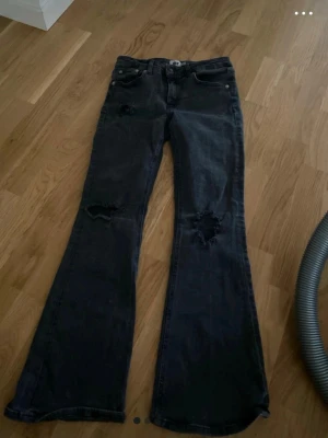 Svarta utsvängda jeans med slitningar - Snygga svarta jeans med utsvängda ben och slitna detaljer på knäna. Jeansen har klassisk femficksdesign och låg midja. Perfekta för en trendig och avslappnad stil.