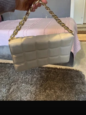 Beige quiltad handväska med kedja - Trendig beige handväska med quiltat mönster och en chunky guldfärgad kedja som axelrem. Väskan har en rektangulär form och är tillverkad i ett slätt, läderliknande material. Perfekt accessoar för att lyfta din outfit.