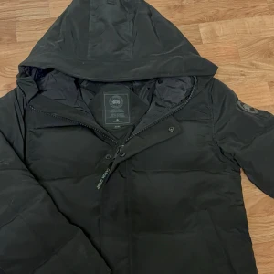 Svart dunjacka från Canada Goose - Snygg svart dunjacka från Canada Goose med huva och dragkedja framtill. Jackan har en klassisk puffer-modell och patch med logga på ärmen. Perfekt för kalla vinterdagar och riktigt varm tack vare dunfyllningen.