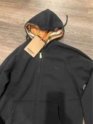 Burberry zip-hoodie  - Svart hoodie från Burberry med klassisk dragkedja och känguruficka. Insidan av huvan har det ikoniska Burberry-rutiga mönstret i beige, rött och svart. Tillverkad i mjukt bomullsmaterial och har en diskret Burberry-logga på bröstet.