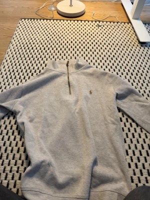 Grå halvzip från Polo Ralph Lauren - Snygg grå halvzip-tröja från Polo Ralph Lauren med broderad logga på bröstet. Tröjan har hög krage och dragkedja framtill, samt ribbade muddar vid ärmslut och nederkant. Perfekt för en stilren och avslappnad look.