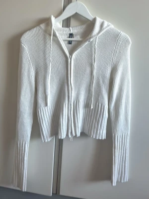 Vit stickad hoodie med dragkedja - Vit stickad hoodie med dragkedja framtill och långa ärmar. Tröjan har ribbstickade detaljer vid ärmslut och nederkant samt en huva med dragsko. Perfekt för en avslappnad och trendig look.