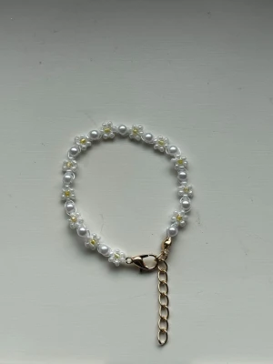 Pärlarmband med små blommor - Handgjort armband med vita pärlor och små blommor i vitt och gult. Armbandet har guldfärgat spänne och justerbar kedja. Perfekt accessoar för att ge en gullig touch till din stil.