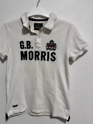 Vit pikétröja från Morris - Vit pikétröja från Morris Gents ett broderat emblem på bröstet. Pikén är gjorde av 100% bomul och modellen har en normal passform. Snygg och stilren. Perfekt för dig som gillar att vara lite stiligare i vardagen