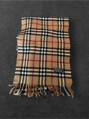 Beige rutmönstrad halsduk Burberry - Klassisk beige halsduk från Burberry med ikoniskt rutmönster i svart, vitt och rött. Halsduken har fransar i ändarna och är tillverkad i mjukt material, perfekt för att ge din outfit en tidlös touch.