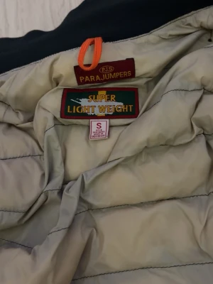 Svart dunjacka från Parajumpers - Snygg svart dunjacka från Parajumpers med gul detalj vid kragen och klassisk logga . Jackan är superlätt och har quiltad design samt dragkedja framtill. Perfekt för kalla vinterdagar och riktigt stilren look. Obs sommarjacka