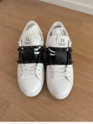 Valentino garavani skor - Unika vita sneakers från Valentino med bred svart rem över vristen dekorerad med svarta nitar. Klassisk låg modell med vita snören och detaljerad logga på plösen. Tillverkade i slätt läder för en clean och edgy look.