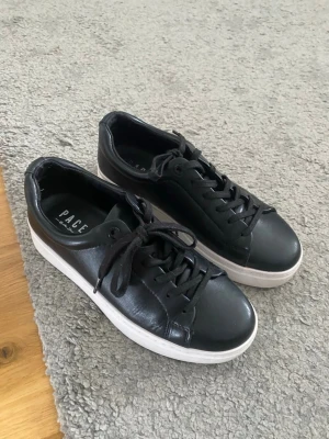 Svarta sneakers från Pace - Stilrena svarta sneakers från Pace med lågt skaft och vita sulor. Skorna har klassisk snörning och är tillverkade i slätt läder för en clean look. Perfekta för dig som gillar minimalistisk stil och vill ha ett par bekväma sneakers till vardags.