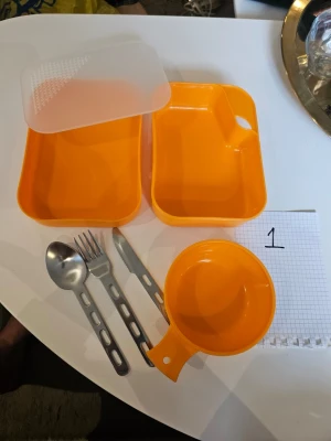 Orange campingmatsats i plast - Praktisk orange campingmatsats i plast med två rektangulära matlådor, lock med sil som även fungerar som skärbräda, skål/kåsa, samt bestickset i metall (sked, gaffel, kniv). Perfekt för utflykter eller friluftsliv, allt får plats i lådan. Säljer 2 set. Detta är set 1