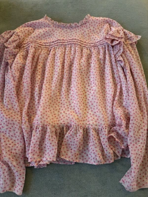 Rosa blommig volangblus - Supersöt rosa blus med små blommor i rött och lila. Blusen har volangdetaljer vid axlar och nederkant, samt långärmade puffärmar. Tunt och luftigt material, perfekt för en romantisk look. Hög hals med liten volangkant.