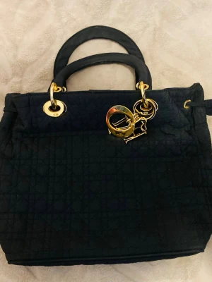 Svart Lady Dior handväska - Säljer en svart Lady Dior handväska i quiltat tyg med guldfärgade metalldetaljer och klassiska Dior-hängen. Väskan har två rundade handtag och en elegant, boxig form. Perfekt för dig som vill ha en ikonisk och stilren accessoar.