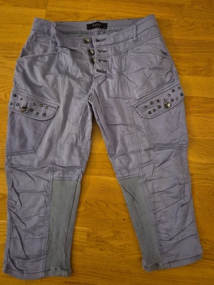 Grå cargobyxor med nitar från PULZ - Snygga grå cargobyxor från PULZ med dekorativa nitar på fickorna och ribbade detaljer vid knäna. Byxorna har flera fickor, knappgylf och en avslappnad passform som ger en cool och trendig look.