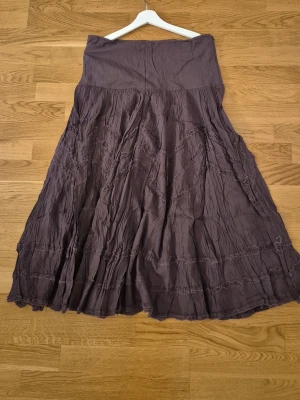 Lång brun kjol med volang - Säljer en lång brun kjol med volangdetaljer och lätt skrynklig look. Kjolen har en bred midja och ett flödande fall som ger en bohemisk känsla. Perfekt för dig som gillar en avslappnad men ändå stilsäker stil.