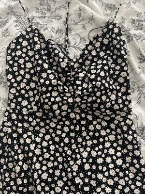 Svart blommig klänning med slits - Söt svart klänning med vita och rosa blommor, smala axelband och en liten slits framtill. Klänningen har en rynkad detalj vid bysten och är i en lätt, luftig modell som passar perfekt till varma dagar.