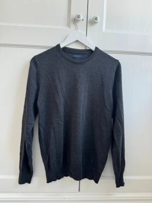 Mörkgrå merinoulls tröja från Berkley - Klassisk mörkgrå stickad tröja från Berkley med rund halsringning och ribbade muddar vid ärmslut och nederkant. Tröjan har en enkel och stilren design, perfekt att matcha med olika outfits.