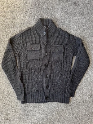 Mörkgrå stickad kofta med knappar - Mörkgrå stickad cardigan från River Island med grovt mönster, kabelstickade detaljer och två bröstfickor med knappar. Koftan har hög krage, axelknappar och långa ärmar. Perfekt för att hålla sig varm och snygg under kyliga dagar.