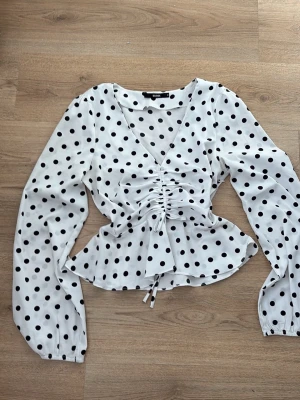 Vit prickig blus med rynkdetalj - Snygg vit blus från BikBok med svarta prickar och trendig rynkdetalj framtill. Blusen har v-ringning, långa ärmar med resår vid ärmslut och knytband i midjan. Tillverkad i ett lätt och luftigt material som ger en skön passform.