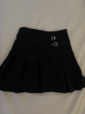 Svart kortkjol med spännen - Säljer en svart kortkjol med två dekorativa spännen i midjan. Kjolen har en lätt utställd form och är perfekt för dig som gillar en edgy och stilren look. Materialet känns mjukt och följsamt, och kjolen har inga synliga mönster eller detaljer förutom spännena. Använt en gång💕storlek 164 men passar mig som vanligtvis har xs/152