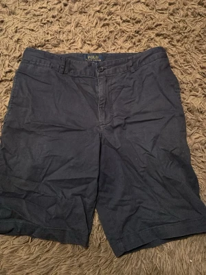 Mörkblå shorts från Polo Ralph Lauren - Snygga mörkblå shorts från Polo Ralph Lauren med klassisk passform. De har bälteshällor, knappgylf och två fickor fram samt två bakfickor med knapp. Perfekta för sommaren och tillverkade i mjuk bomull.