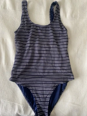 Randig marinblå baddräkt - En klassisk baddräkt i marinblått med vita horisontella ränder. Modellen har breda axelband och rundad urringning både fram och bak. Tillverkad i stretchigt material som sitter bekvämt på kroppen. Perfekt för stranden eller poolen.