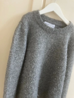 Grå stickad tröja från Bershka - Mysig grå stickad tröja från Bershka med rund halsringning och ribbade muddar vid ärmslut och nederkant. Tröjan har en enkel, rak passform och är tillverkad i ett mjukt, fluffigt material som passar perfekt för kyligare dagar, och bara ett vanligt basic vardagsplag.