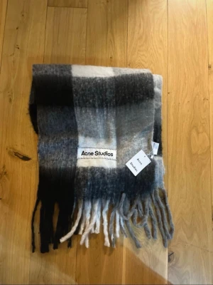  Acne Studios halsduk - Säljer en fluffig och mjuk halsduk från Acne Studios i rutigt mönster med nyanser av svart, grått och vitt. Halsduken har långa fransar i ändarna och en stor vävd logotyp. Perfekt accessoar för att lyfta din outfit.