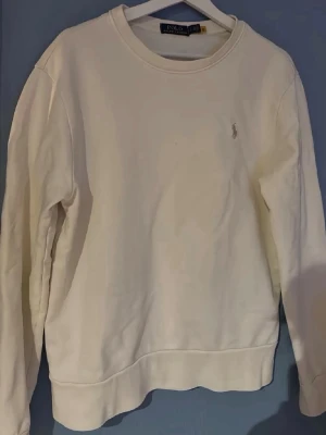 Vit sweatshirt från Polo Ralph Lauren - Klassisk vit sweatshirt från Polo Ralph Lauren med rund halsringning och diskret broderad logga på bröstet. Tröjan har ribbade muddar vid ärmslut och nederkant, perfekt för en clean och stilren look.