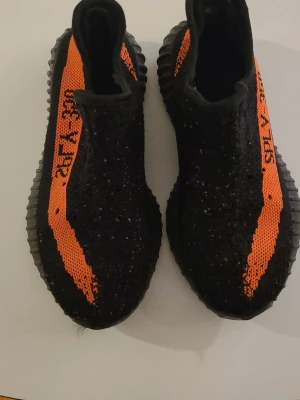 Yeezy Boost 350 V2 Black/Orange - Säljer ett par Yeezy Boost 350 V2 med svart Primeknit-överdel och orange sidostrip med texten 'SPLY-350'. Skorna har en chunky, räfflad sula i svart och den ikoniska silhuetten som är superpopulär bland sneakerheads.