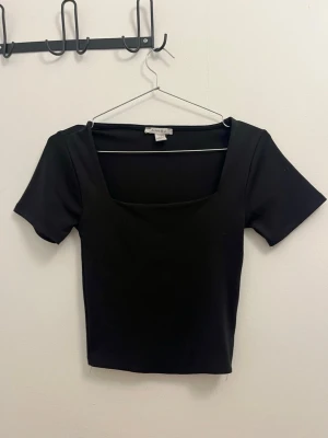 Svart croppad topp från Ambisu - Snygg svart croppad topp från New Yorker med fyrkantig ringning och korta ärmar. Perfekt till höga jeans eller kjol. Enkel och stilren design som passar till många olika looks.