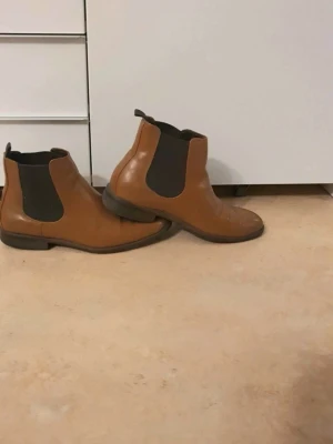Bruna Chelsea boots  - Snygga bruna Chelsea boots med klassiska svarta resårpaneler på sidorna och låg klack. Perfekta för en stilren look och lätta att ta på tack vare resåren. Passar till både jeans och kostymbyxor.