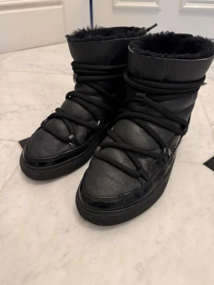 Svarta Inuikii moon boots - Säljer ett par svarta moon boots från Inuikii med högt skaft och snörning runt om. Bootsens utsida är i glansigt och matt läder, och insidan är fodrad med mjuk svart pälsimitation. Perfekta för kalla dagar och riktigt snygga till vinterlooken.