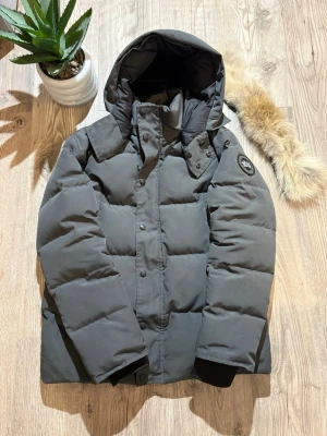 Canada Goose - Snygg mörkgrå dunjacka från Canada Goose med avtagbar huva och patch-logga på ärmen. Jackan har stora quiltade partier, hög krage och tryckknappar framtill. Perfekt för kalla vinterdagar och riktigt varm.